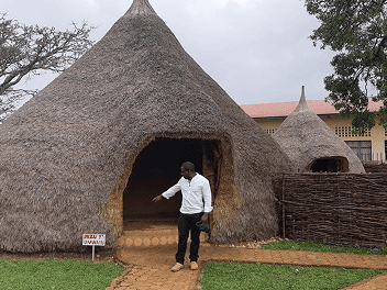 Imagem de um homem de camisa social branca e calça preta, à porta de uma cabana e apontando para uma placa no canto direito da entrada dessa cabana